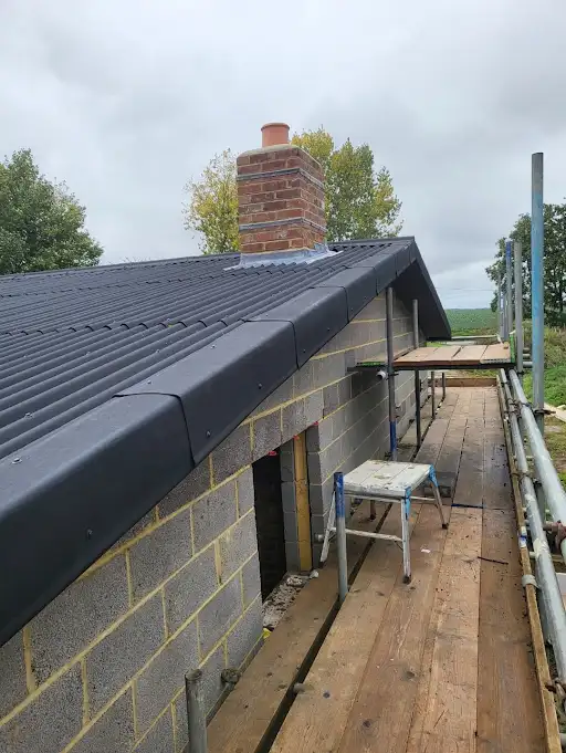 Scaffolding alongside a building with gray corrugated roof and brick chimney under a cloudy sky.