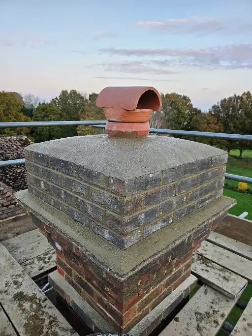 Brick chimney with cement top and terracotta chimney pot, surrounded by scaffolding and trees in background.