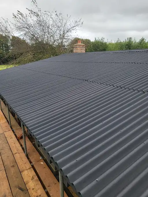 Newly installed dark gray corrugated metal roof viewed from above with trees and chimney in background.