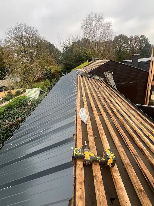Partially installed metal roof panels next to wooden roof battens with drill on top, outdoors under cloudy sky.