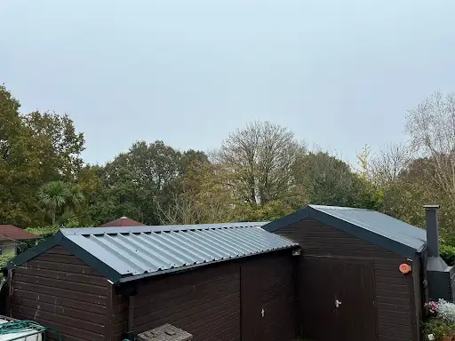 Two connected dark wooden garden sheds with corrugated metal roofs surrounded by trees.