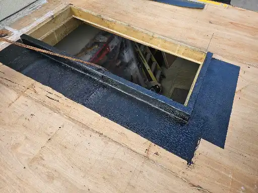 Open rectangular floor hatch with black sealant around wooden frame revealing a lower level space.