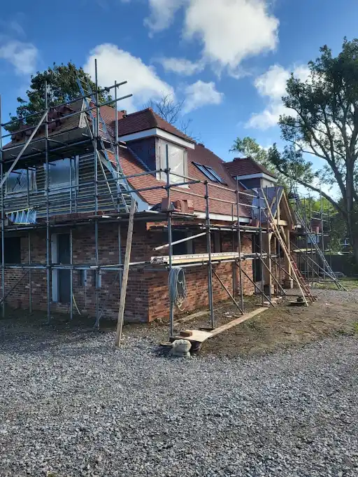 Brick house under construction with scaffolding around it on a gravel yard under blue sky.