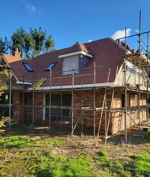 Brick house under construction with scaffolding and a red tiled roof against a blue sky.