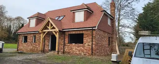 New brick house under construction with red roof and dormer windows in a rural area.