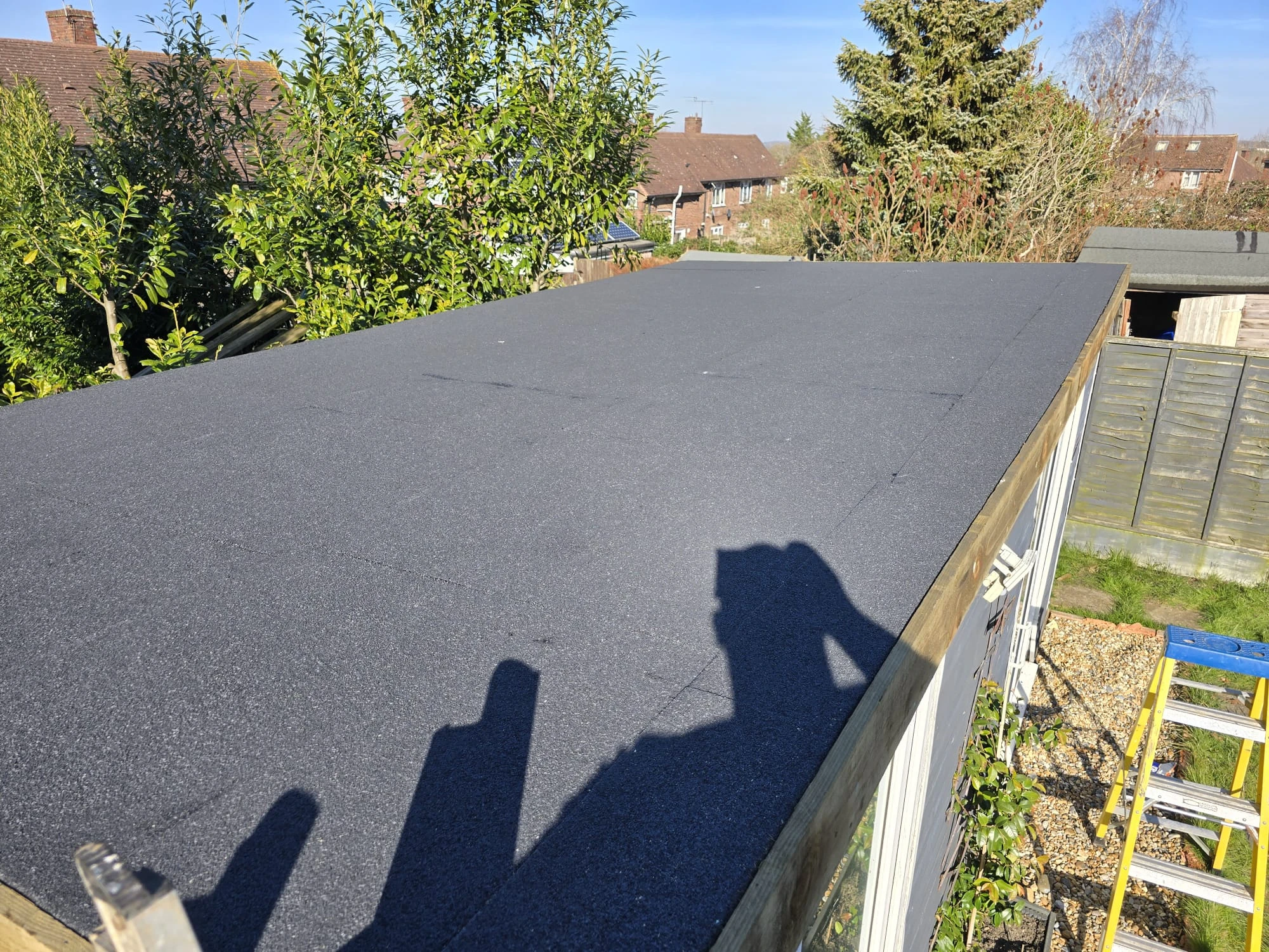 Flat black roof on a wooden structure with trees and houses in the background under a clear sky.